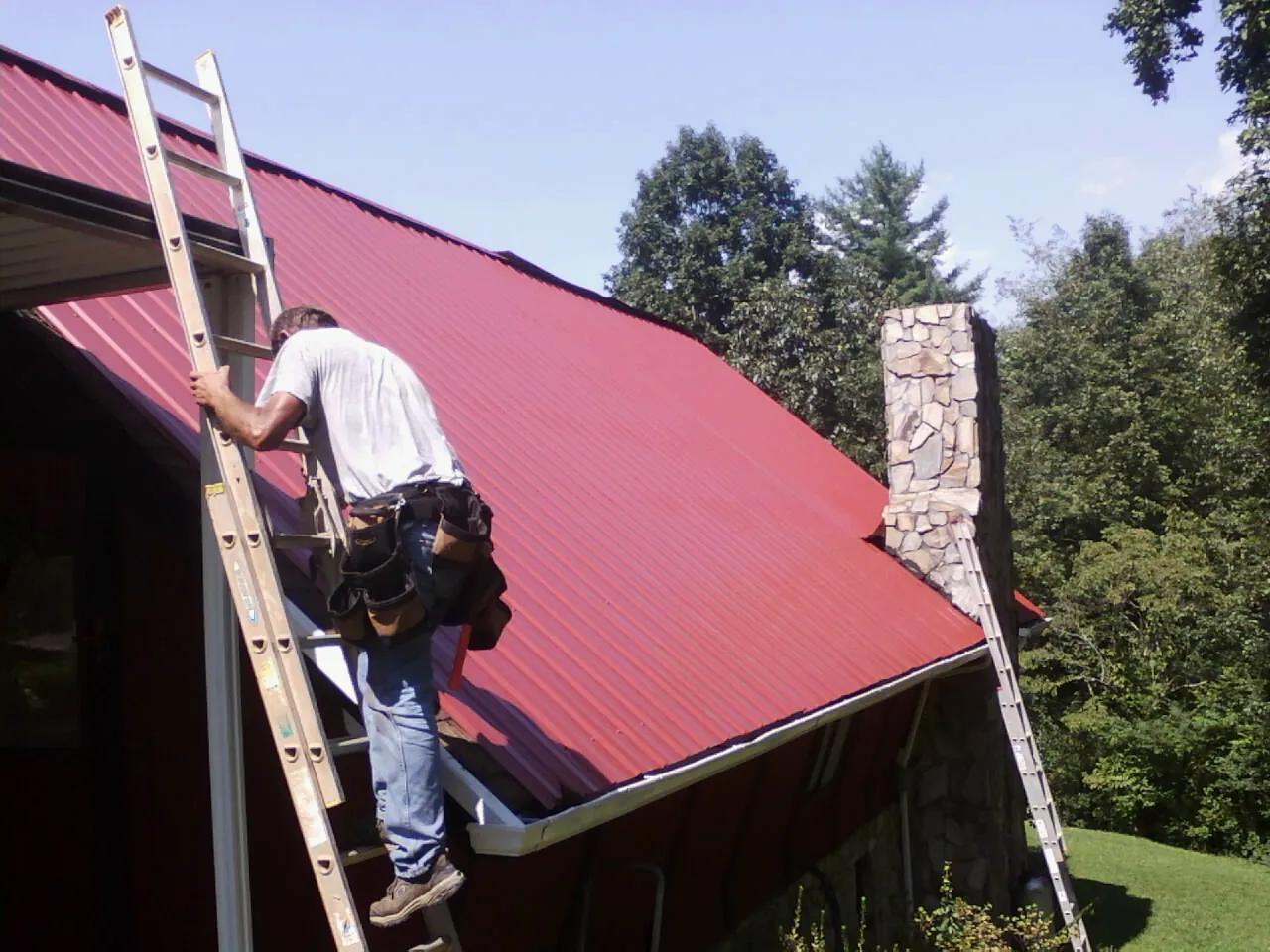 Roofer on a ladder beside a red metal roof and stone chimney — Soffit & Fascia Repair by Roofing Brothers in Manitou